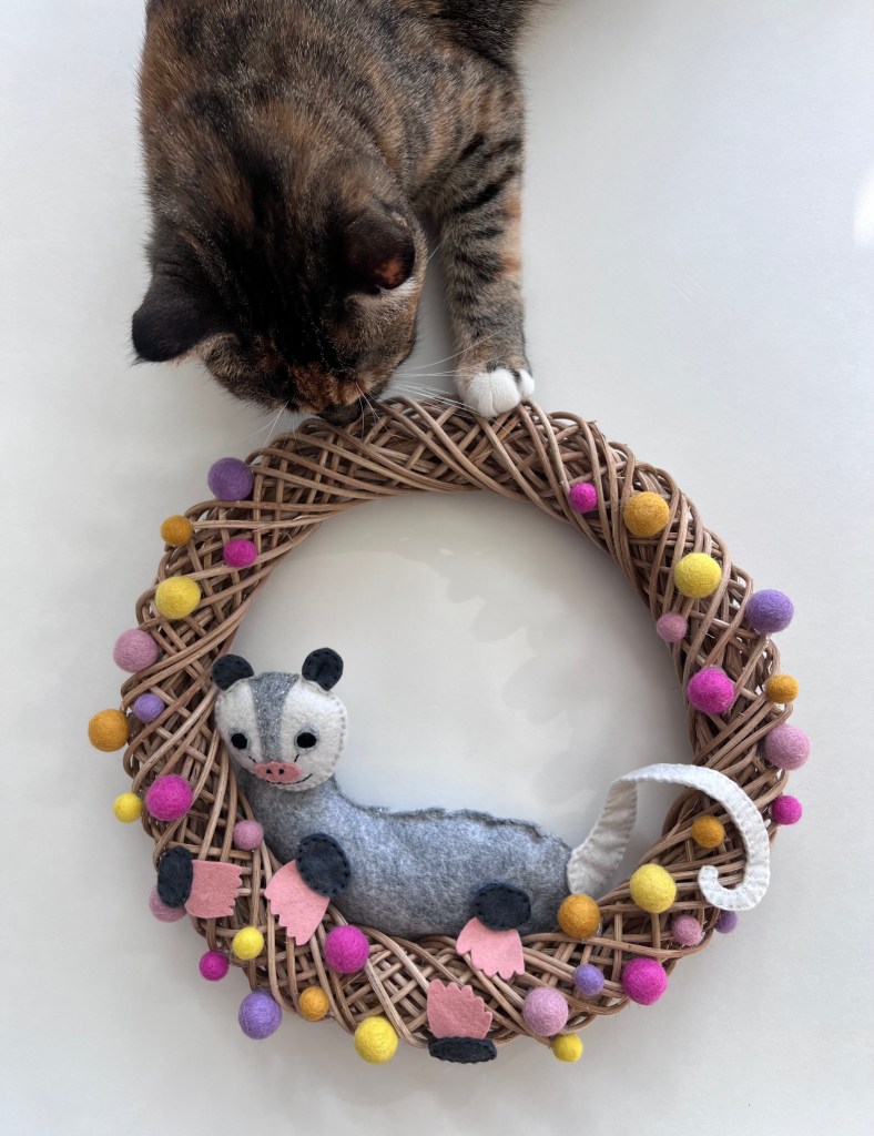 A cat playfully reaches out to a wreath featuring a felt opossum and Pom poms.