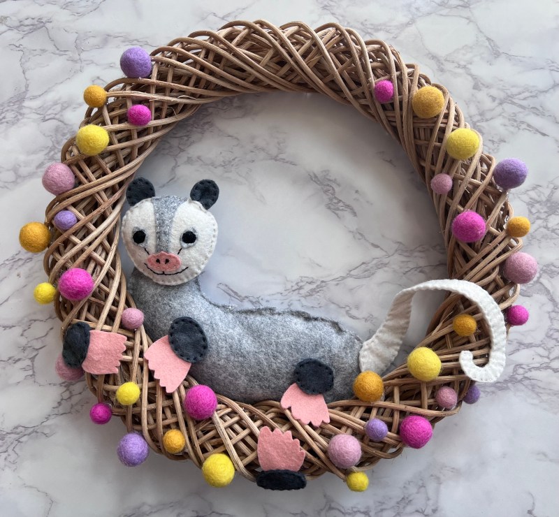 A vine wreath with an opossum made of felt sitting inside and decorated with pastel Pom poms. 